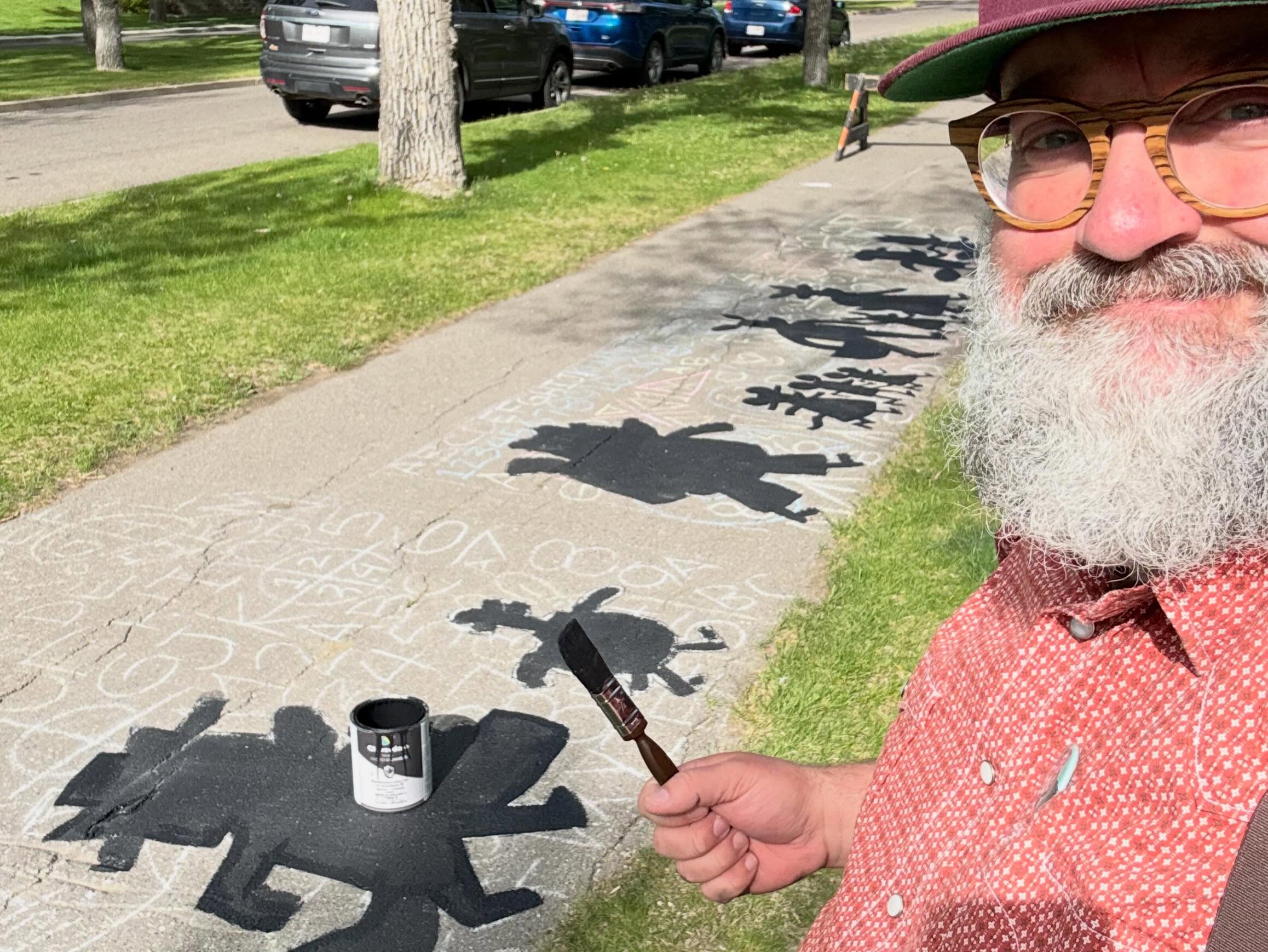 A man with a beard and a paintbrush whimsically gestures in front on an in progress painting on the park pathway. 