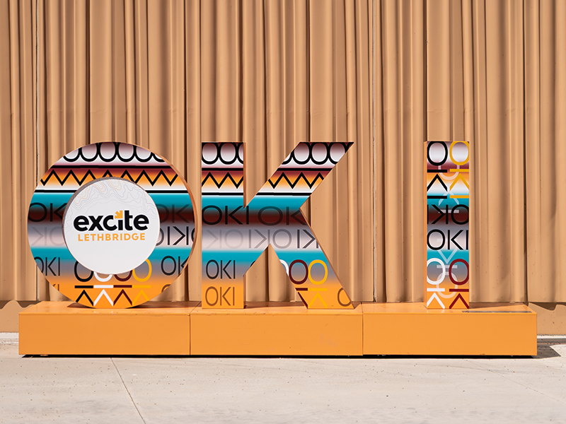 OKI by Cheyenne (Naatoiyiki) McGinnis OKI Sculpture inside at Excite Lethbridge by Cheyenne (Naatoiyiki) McGinnis. The design across the three letter plays with word OKI on a trade-blanket inspired background using golds, browns and turquoise colours.