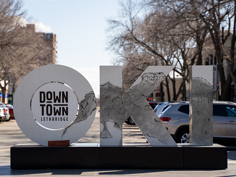 OKI by Wayne Provost Naatosiohpaipiiyi – Sunjumer OKI sculpture by Piikani artist – Wayne Provost Naatosiohpaipiiyi – Sunjumer in Festival Square in downtown Lethbridge with a smokey, charcoal landscape of the coulees across the three letters.